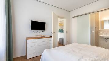 Ein Schlafzimmer mit Bett, Kommode, Fernseher und einem Badezimmer mit Waschbecken und Spiegel.