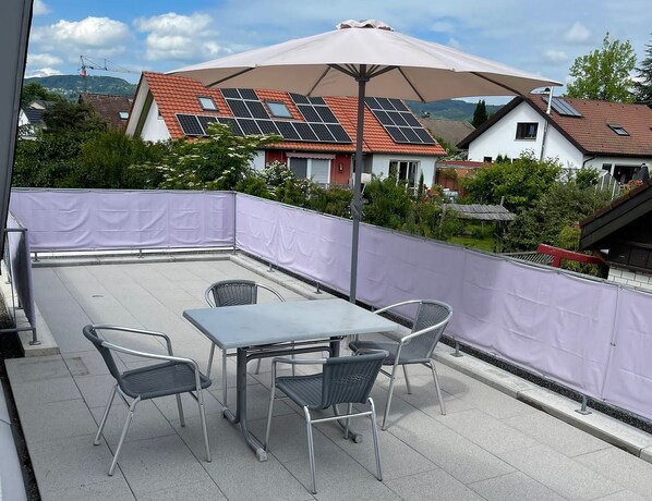 Un patio en la azotea con una mesa y sillas, paneles solares en el techo y vistas a casas al fondo.