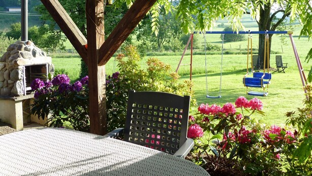 A covered patio with a stone fireplace, a swing set, and a garden with flowers.