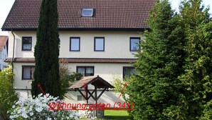 A two-story house with a tiled roof, a small attached structure, and a well-maintained garden with white flowers.