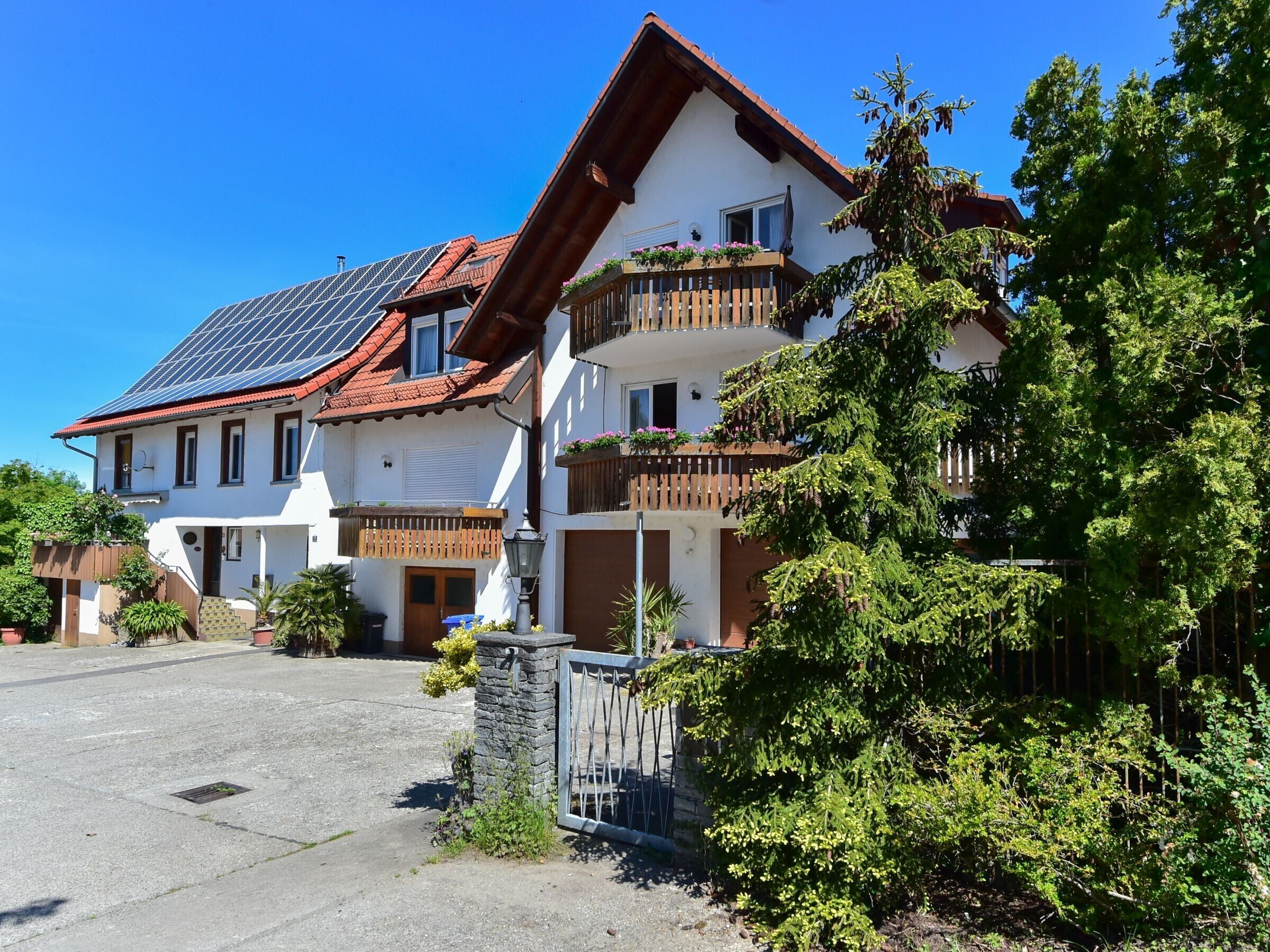 Ein zweistöckiges Gebäude mit einem geneigten Dach, Solaranlagen und einem Balkon mit Blumentöpfen.