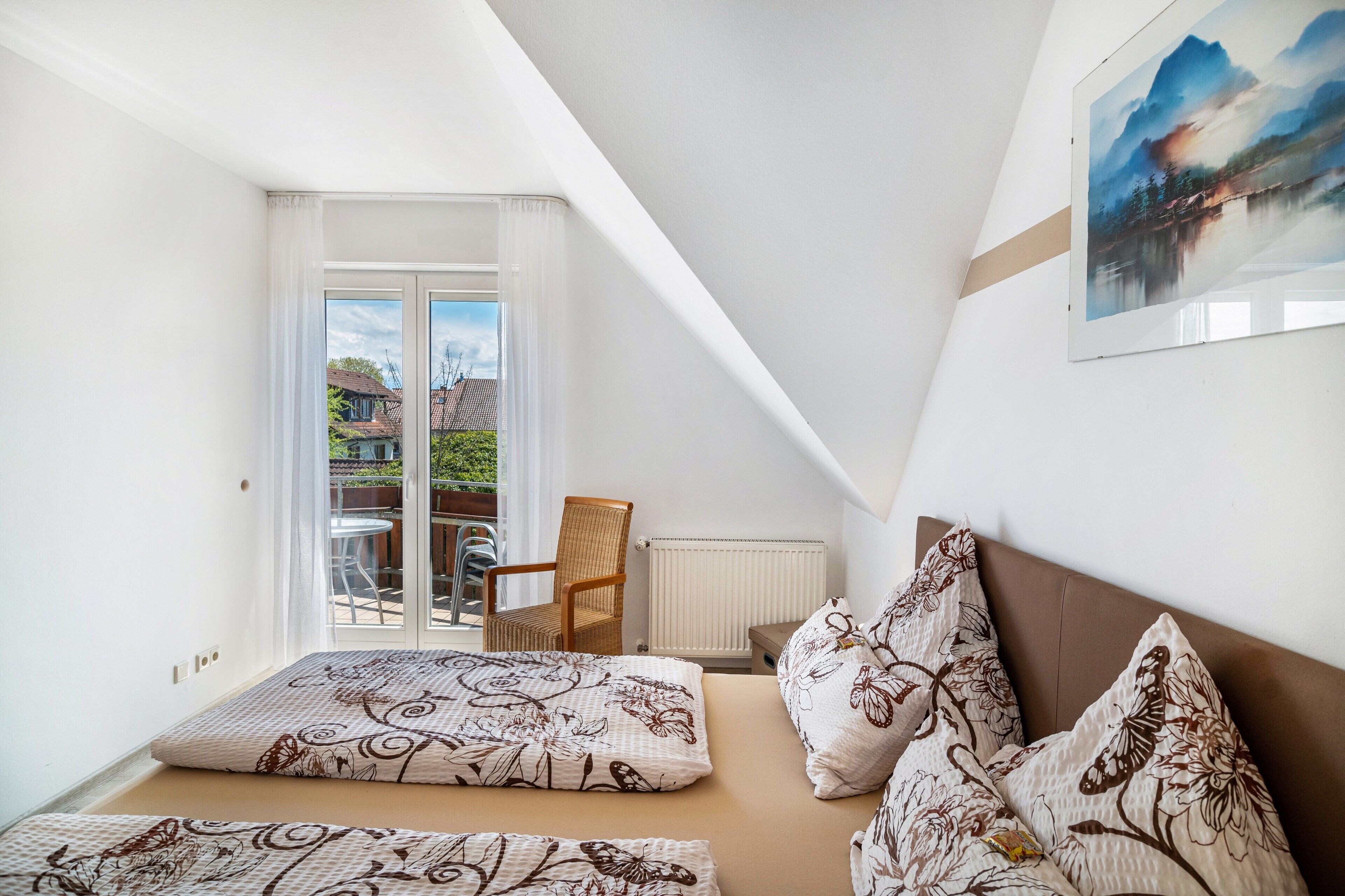 A bedroom with a bed, pillows, a chair, a table, and a view of the outside.