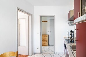 Une cuisine avec un sol carrelé, des placards en bois et une porte visible donnant sur une autre pièce.