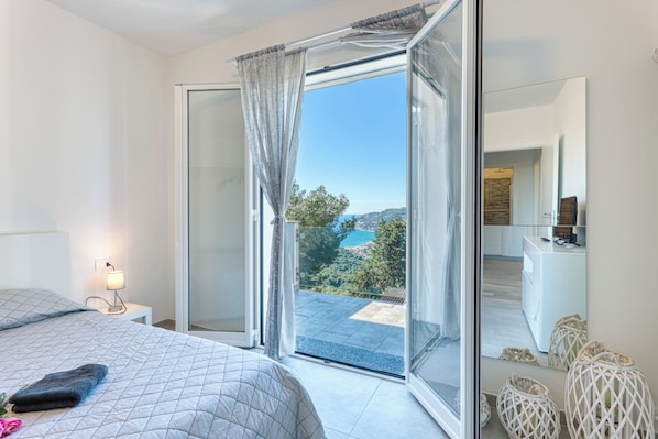 A bedroom with a bed, a nightstand with a lamp, a sliding glass door to a balcony, and a view of a landscape.