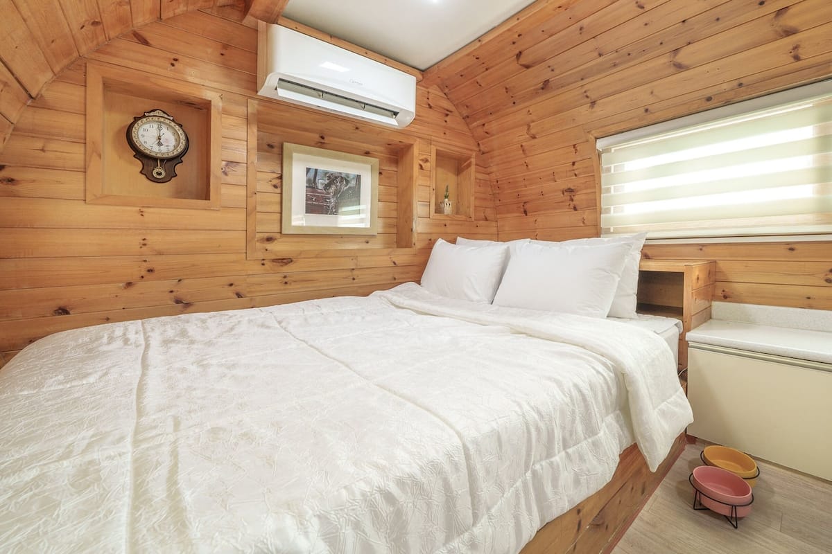 A wooden cabin bedroom with a bed, a clock, an air conditioner, a framed picture, and a window with blinds.
