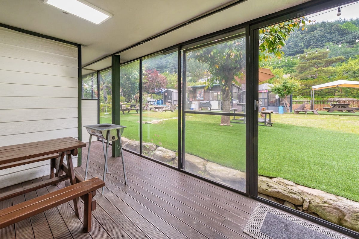 A covered patio with a wooden bench and a grill, large glass windows offering a view of a grassy yard with picnic tables and a building in the background.