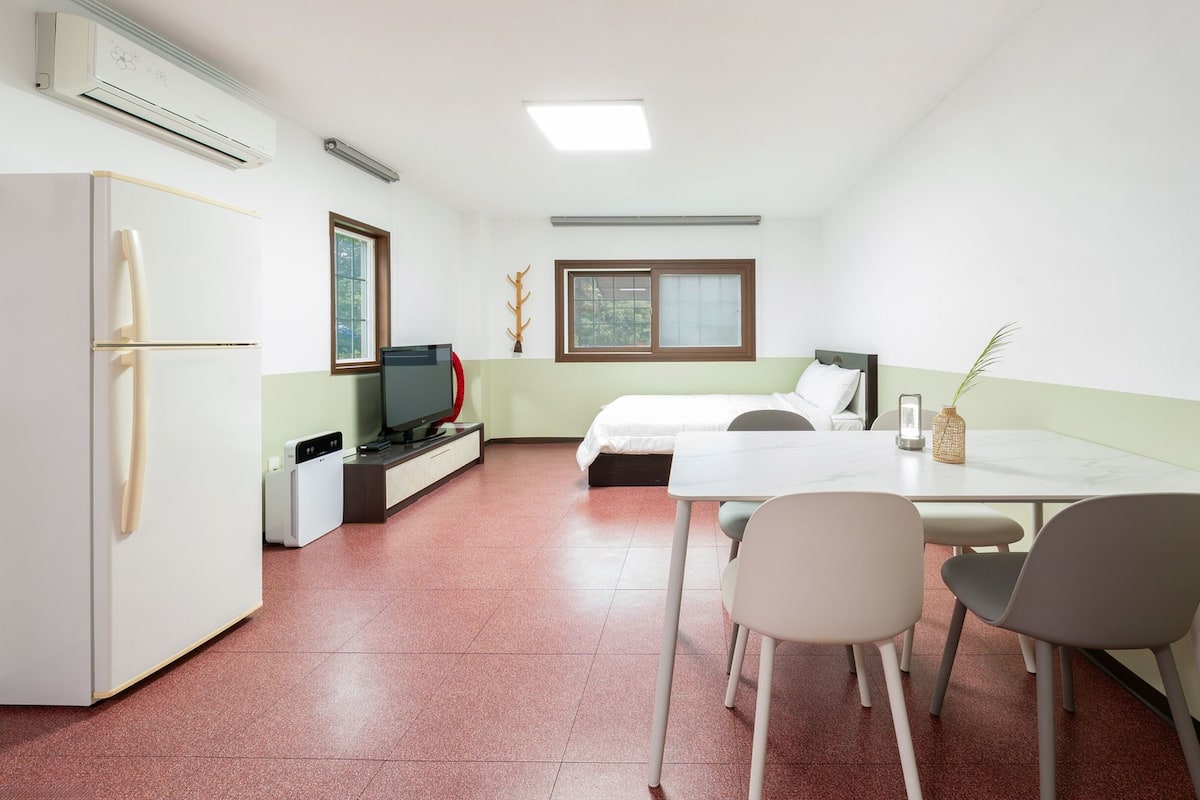 A compact hotel room with a bed, a desk with a chair, a small kitchenette, and a TV.