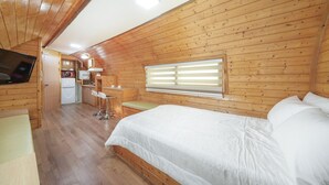 A wooden cabin interior with a bed, a small kitchenette, a TV, and a window with blinds.