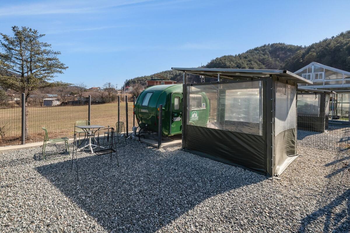 Un pequeño espacio exterior cerrado con una caravana verde, una zona de estar cubierta y suelo de grava.