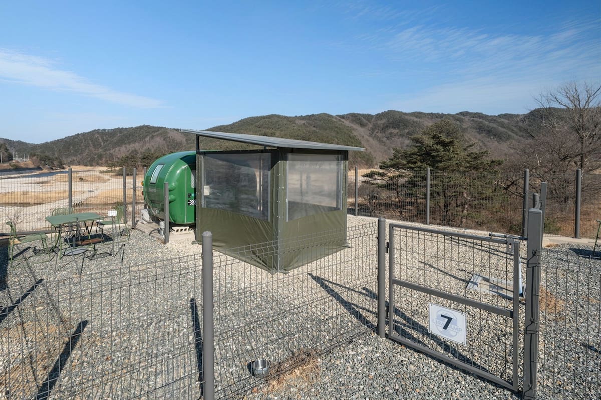 Un área exterior cercada con un baño químico verde, una mesa de metal con sillas y un refugio.