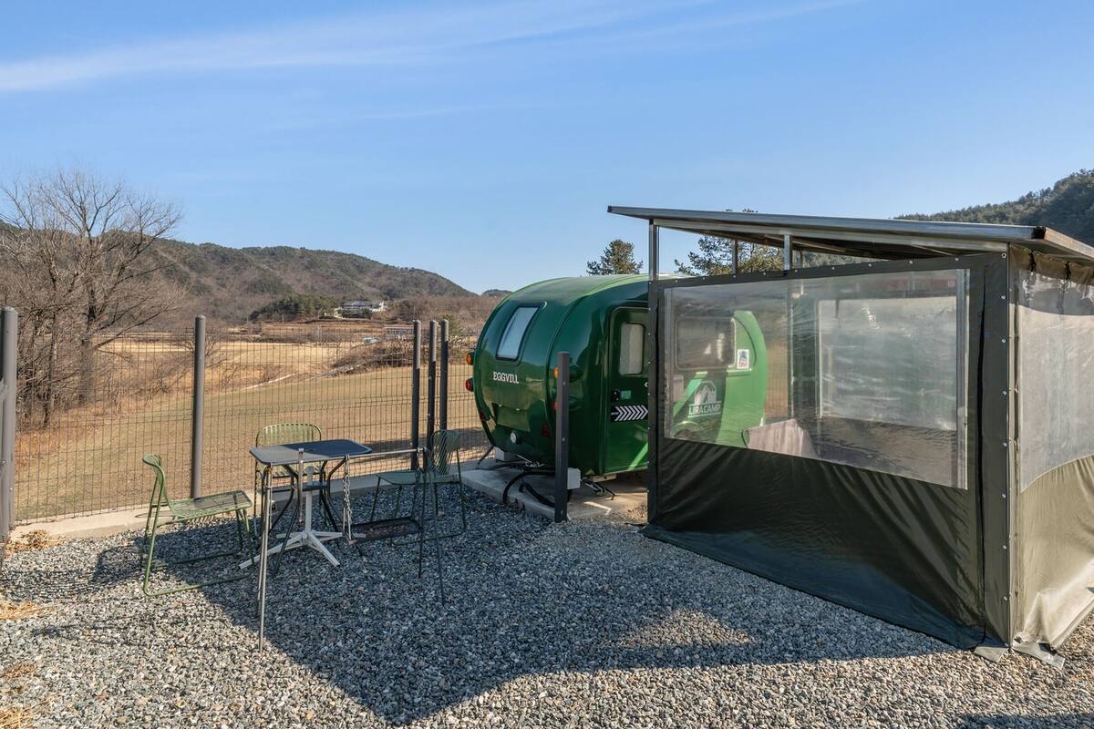 Una autocaravana verde con un área cubierta, una mesa y sillas en un espacio con grava, una cerca y colinas al fondo.