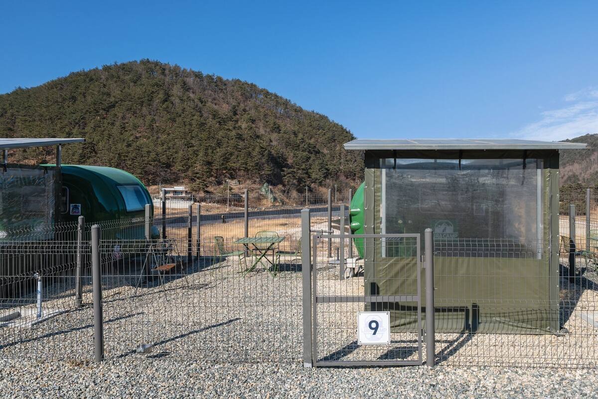 Un baño portátil con valla metálica y un letrero con el número 9, con un cerro boscoso y un cielo despejado de fondo.