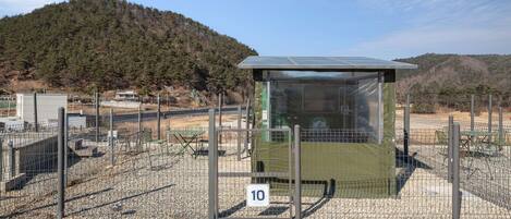 A small, enclosed structure with a metal roof, surrounded by a wire fence, set against a backdrop of a forested hill.