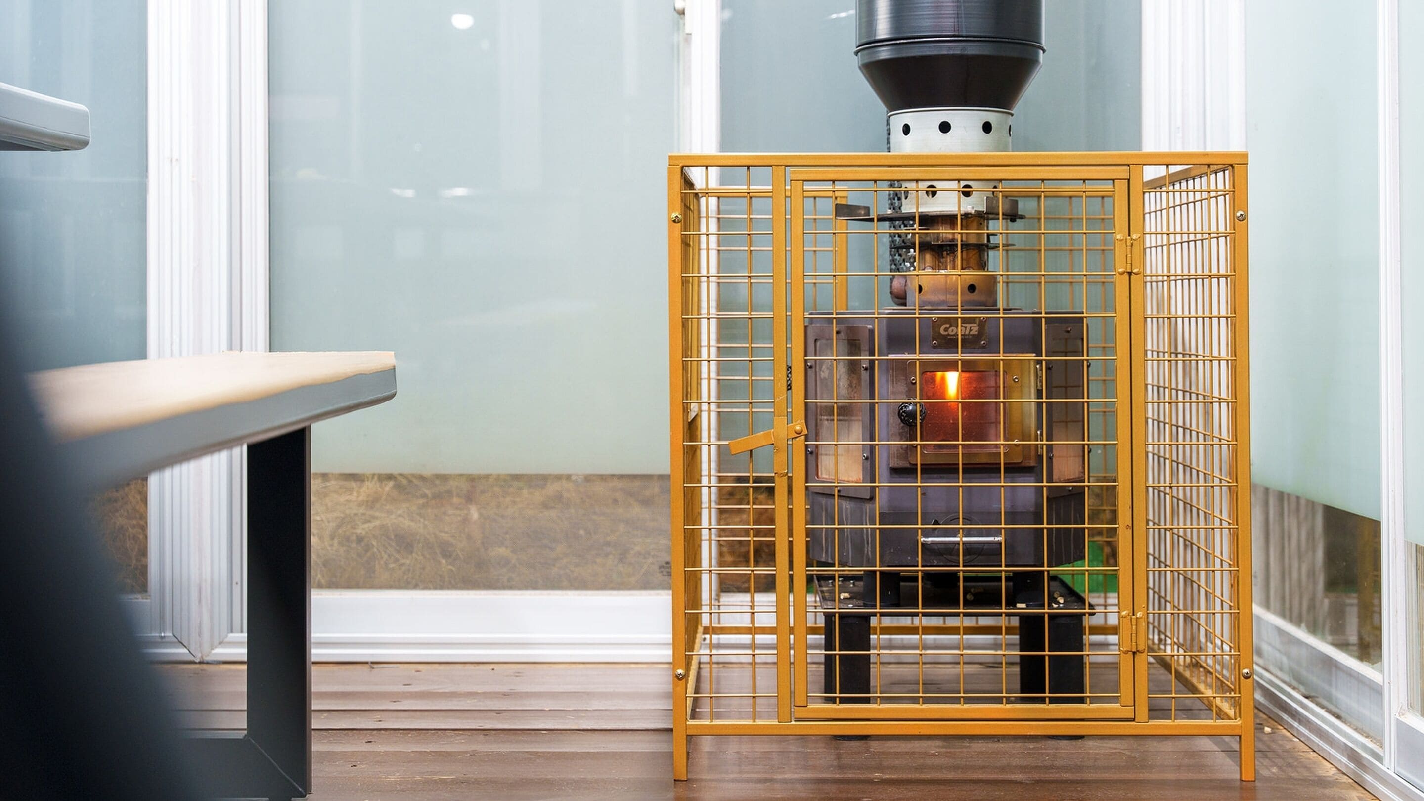 A wood-burning stove encased in a yellow metal grill, with a visible chimney and a window in the background.