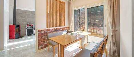 A modern living room with a wooden table and chairs, a fireplace, and a large window with a view of trees.