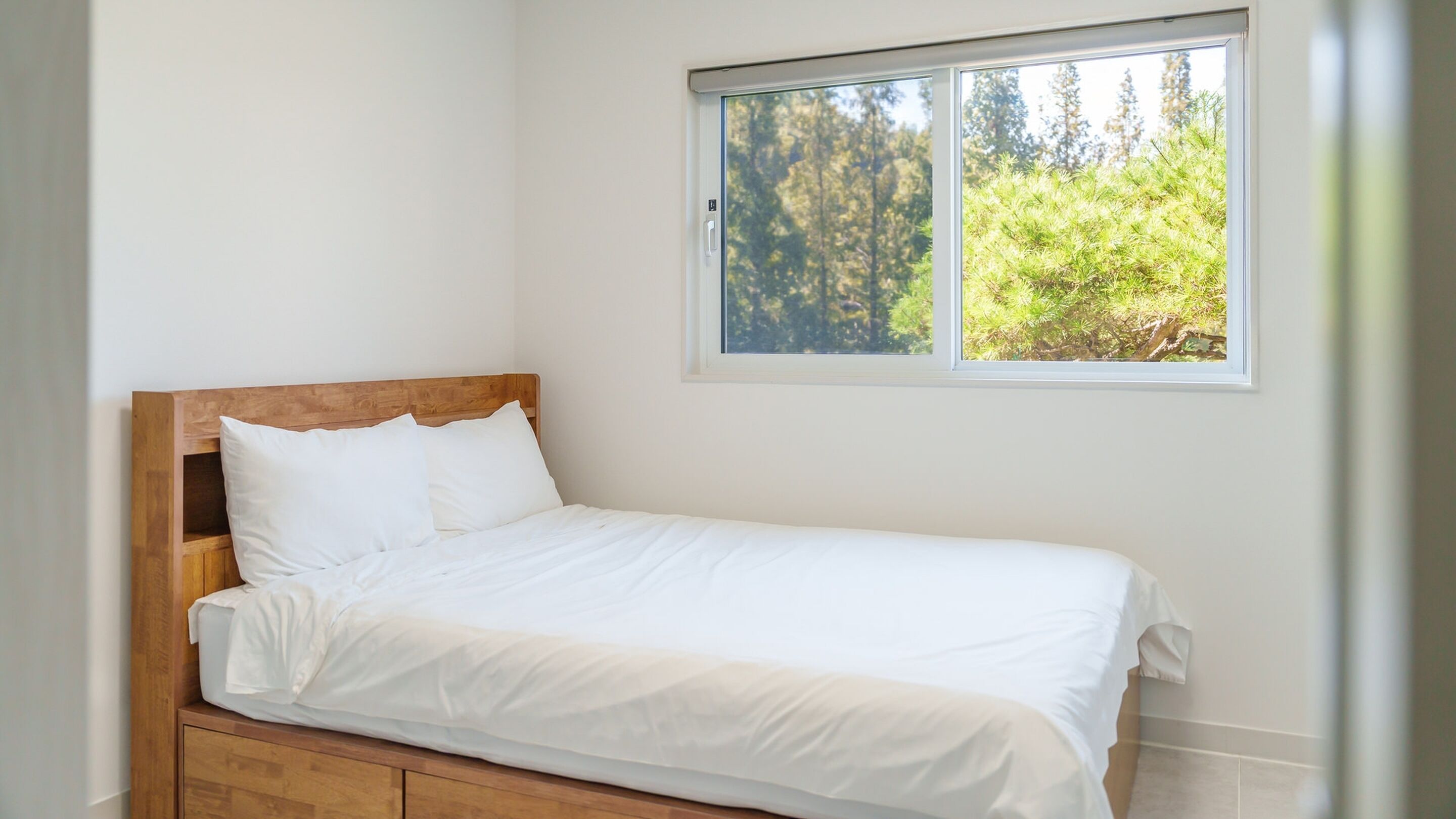 A single bed with white bedding in a room with a window overlooking a forest.