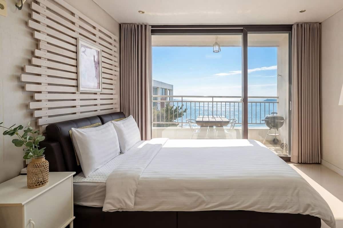 Un dormitorio con cama, mesita de noche, lámpara, vista al mar y balcón con mesa y parrilla.