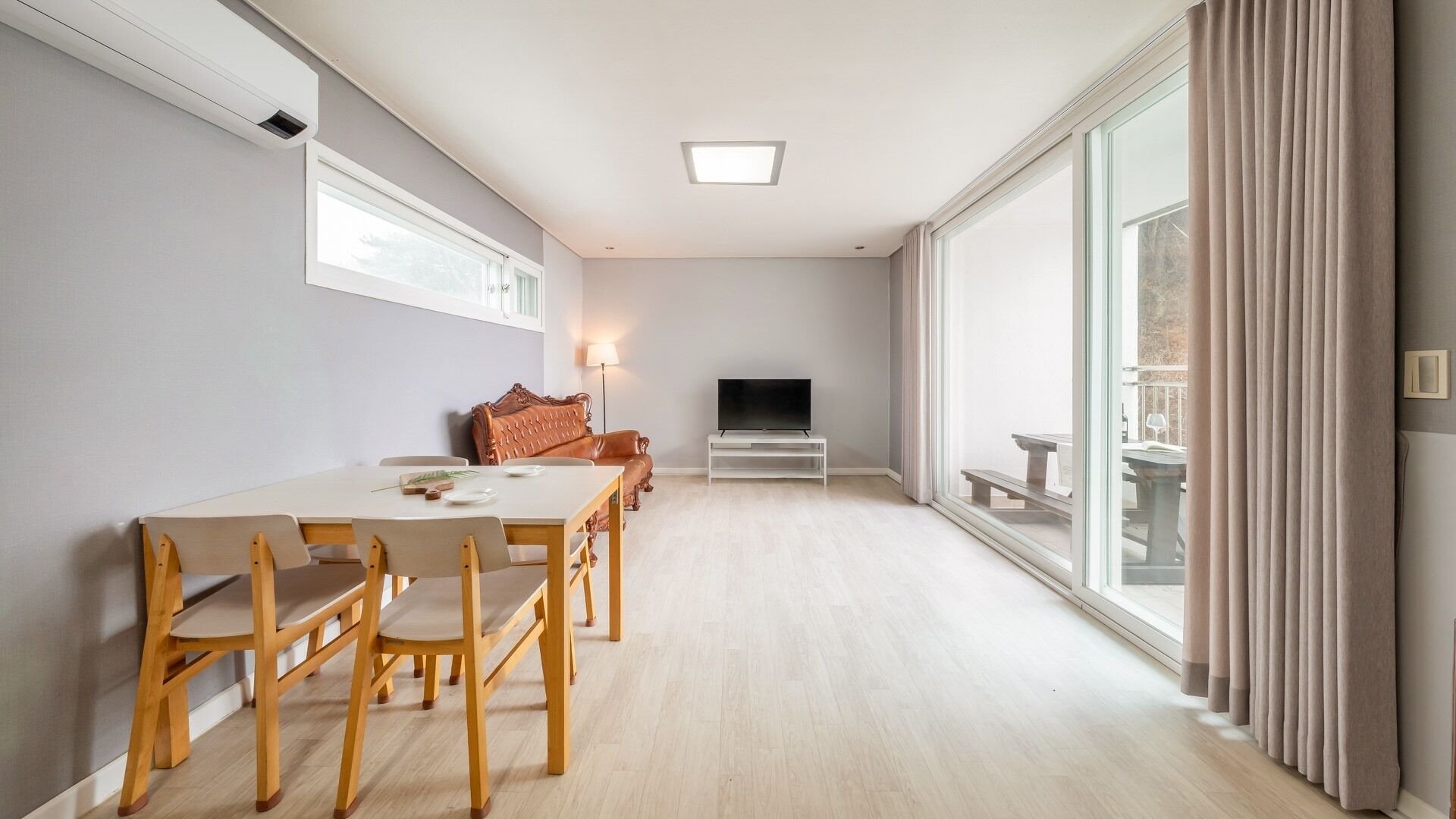 A modern living room with a dining area, a flat-screen TV, and a balcony with curtains.