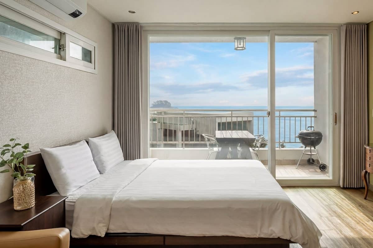A hotel room with a bed, a desk, a TV, a lamp, and a balcony with a view of the sea.