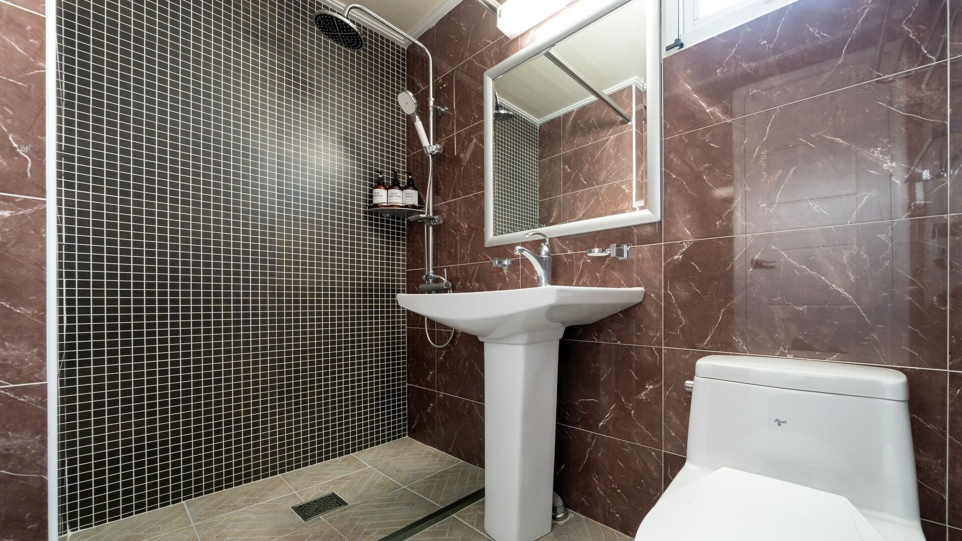 A bathroom with a shower, sink, and toilet, tiled with dark and light tiles.