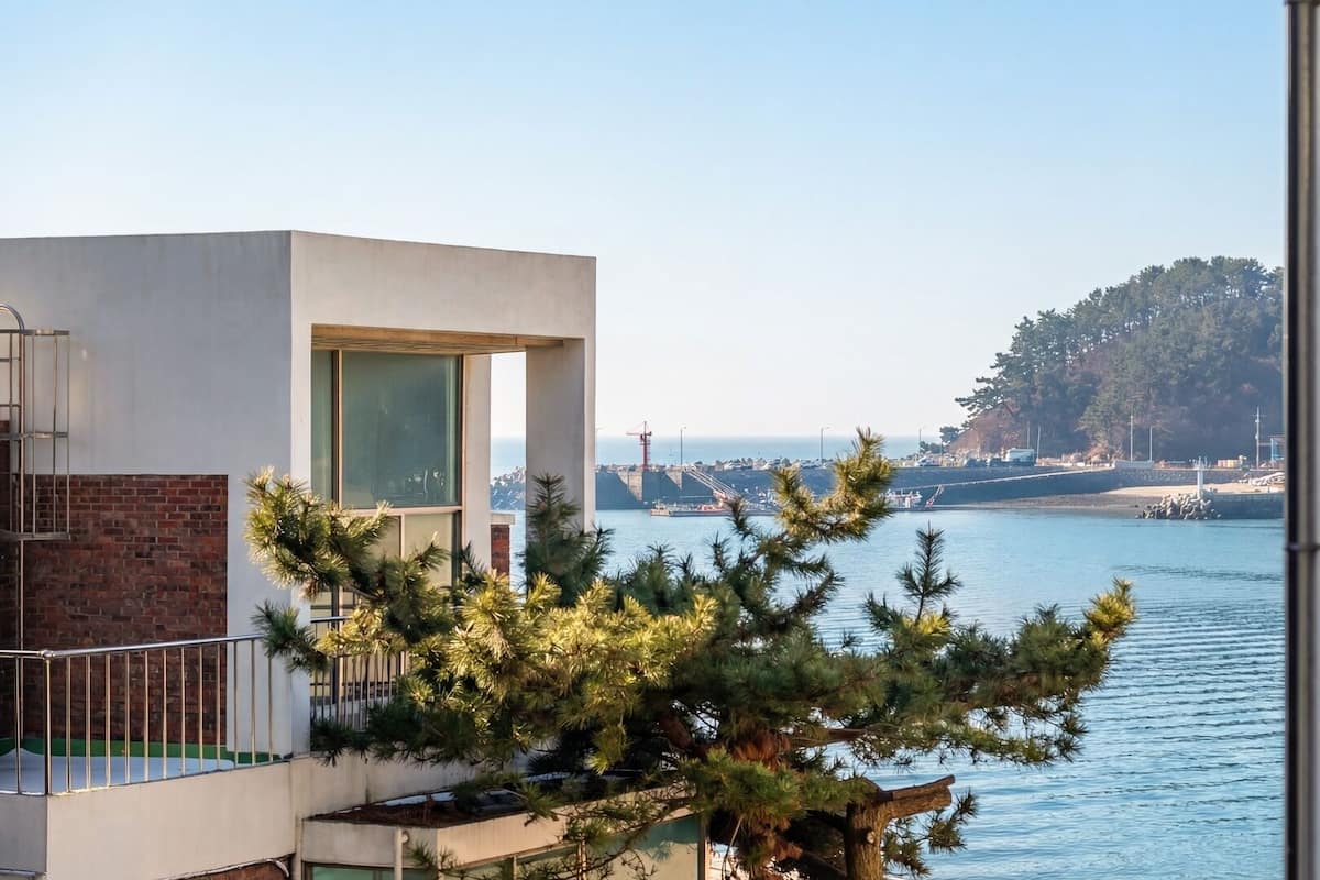 A modern building with a balcony overlooking a calm body of water, a distant tree line, and a clear sky.