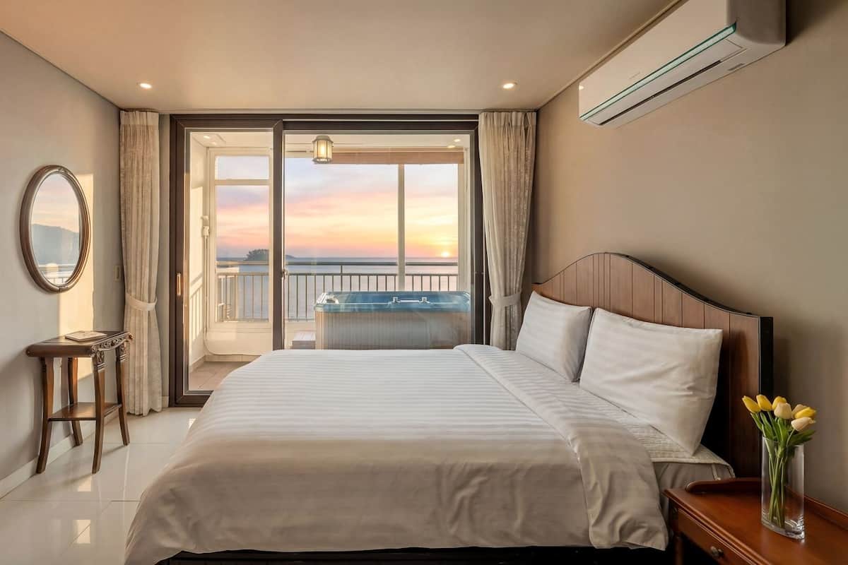 A hotel room with a bed, a desk, a lamp, a mirror, a vase with flowers, and a view of the sunset through the balcony doors.