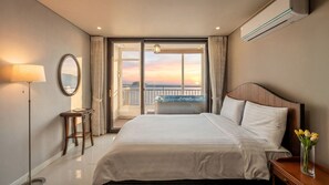 Habitación de hotel con cama, escritorio, lámpara, espejo, un jarrón con flores y vista al atardecer a través de las puertas del balcón.