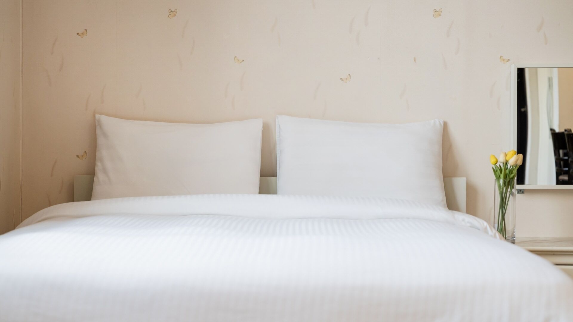 A neatly made bed with white linens, a wall with a subtle pattern, and a vase with flowers on a nightstand.
