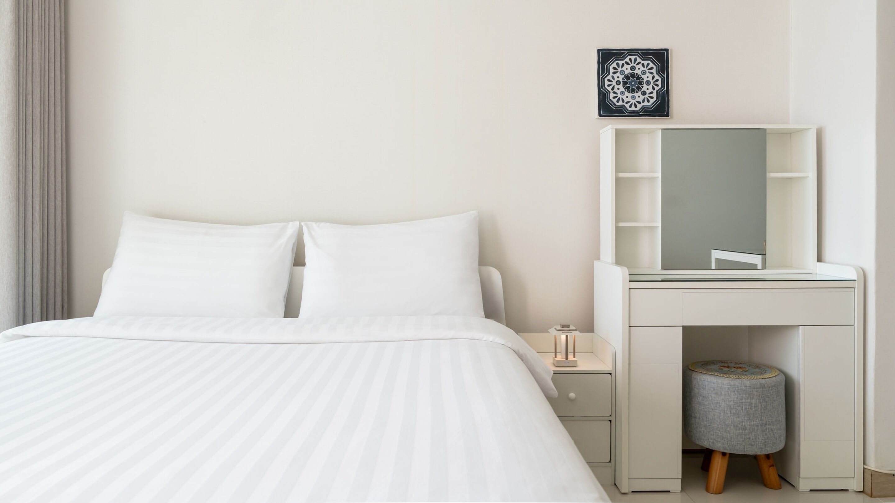 A hotel room with a bed, a white dresser with a mirror, a small stool, and a decorative wall piece.