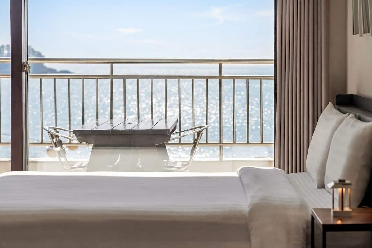 Habitación de hotel con cama, mesita de noche, mesa y sillas, y balcón con vistas.