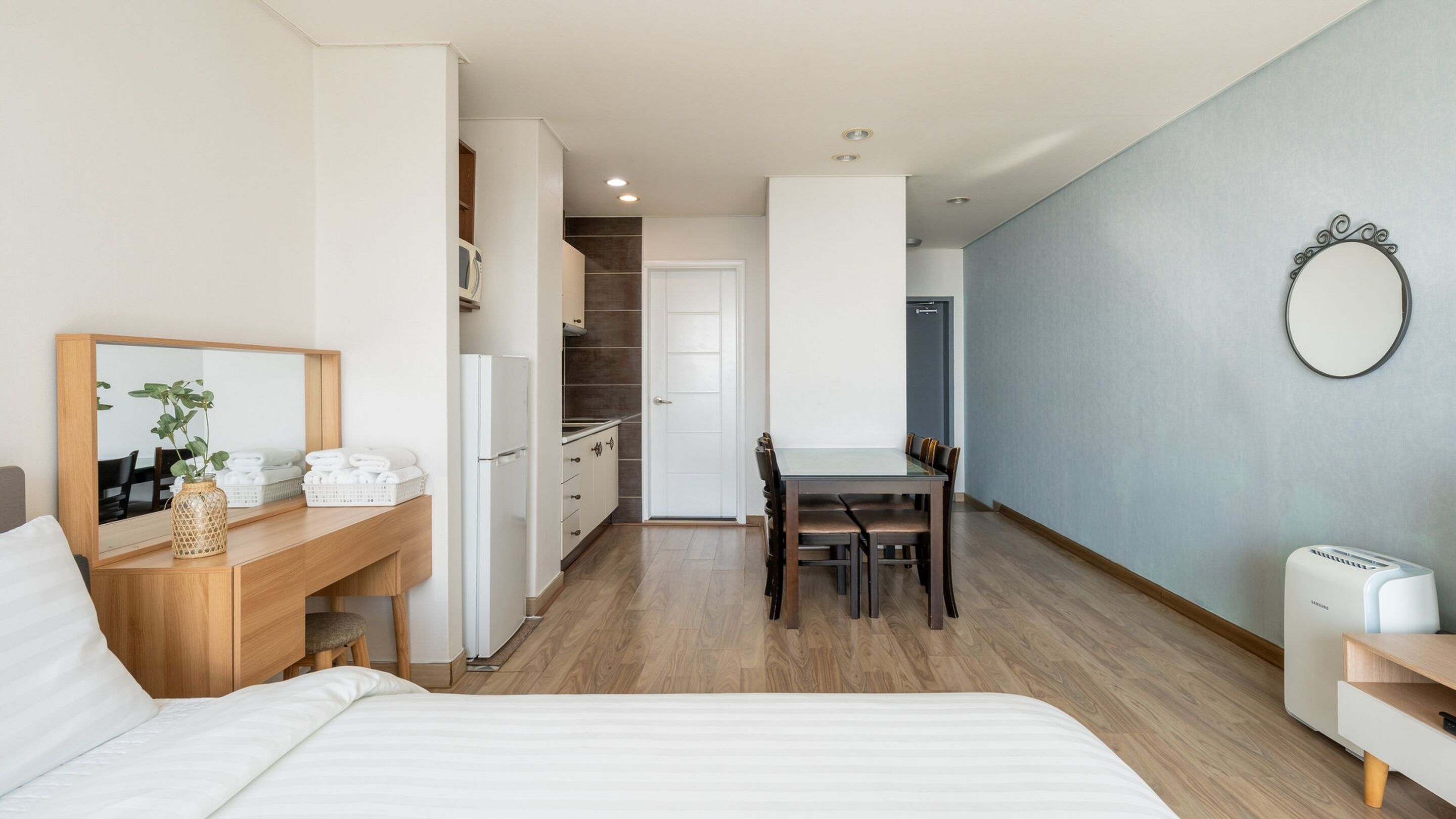Una habitación de hotel moderna con cama, escritorio con espejo, mesa de comedor con sillas, zona de cocina y baño.
