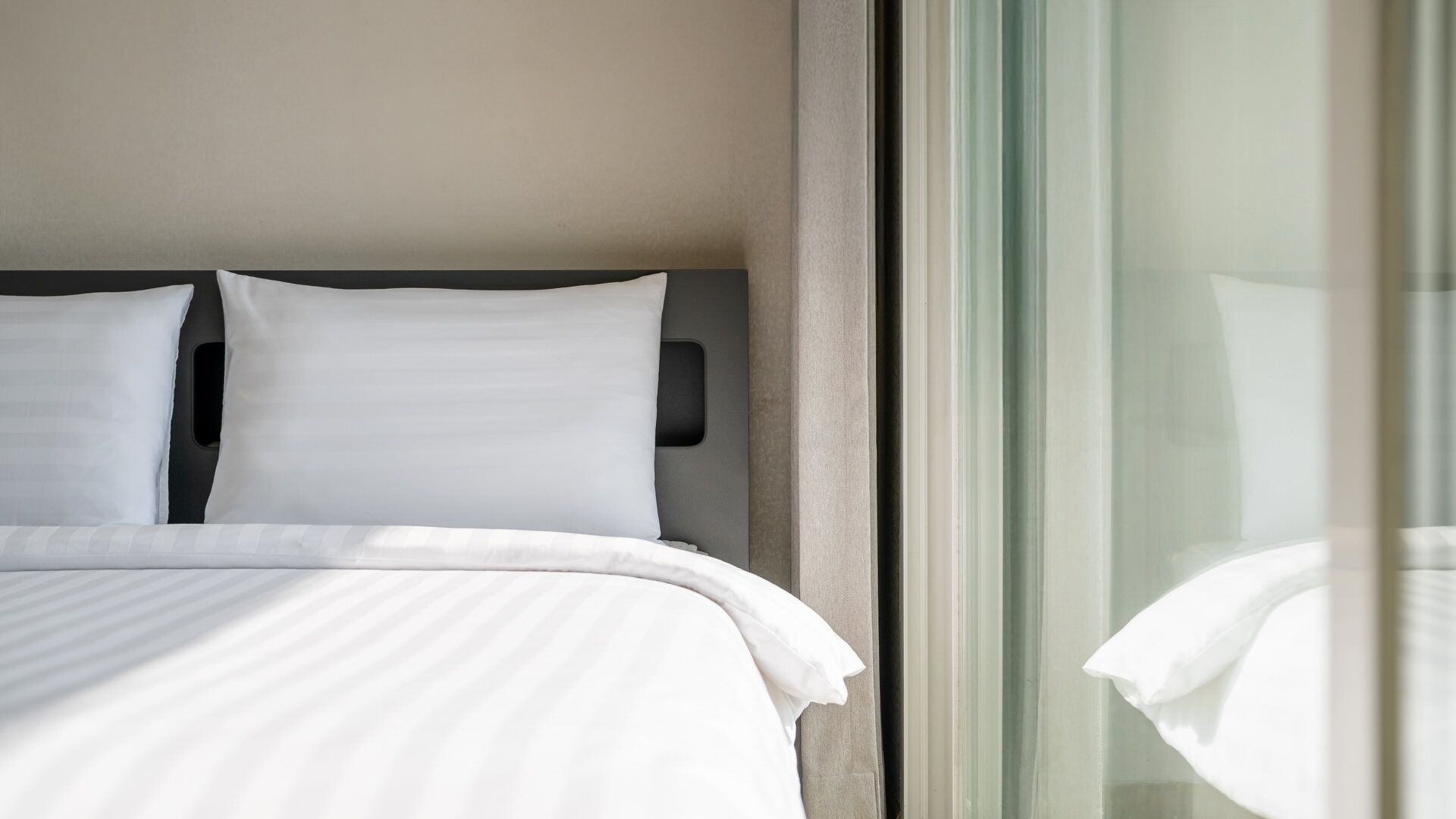 Ein Bett mit weißer Bettwäsche und Kopfteil, ein Fenster mit Vorhängen und Blick auf ein Gebäude.