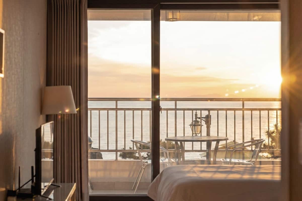 Habitación de hotel con un ventanal grande, una cama, una mesa con sillas y una lámpara.