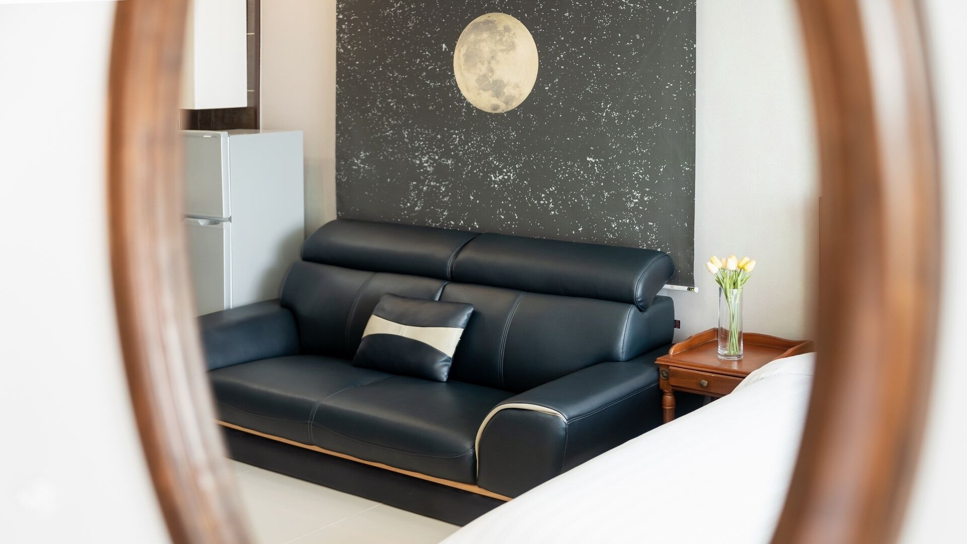 A modern room with a dark leather sofa, a small wooden side table with a vase of flowers, and a wall with a circular moon-like design.