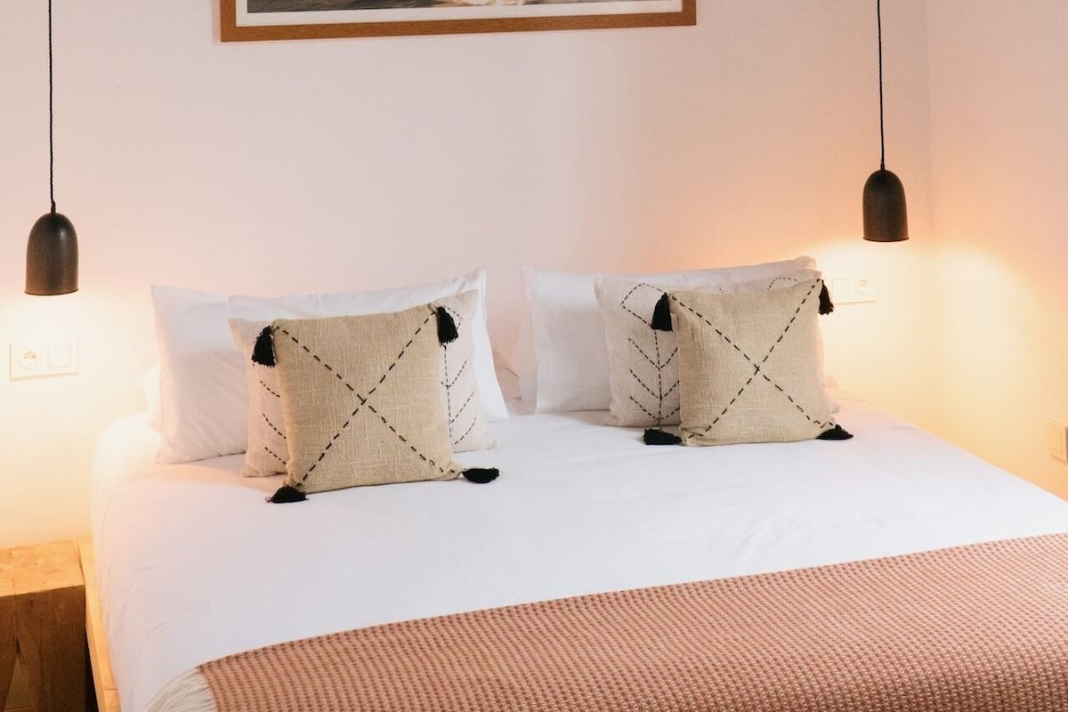 A neatly made bed with white linens, two pillows with a leaf pattern, and a woven pendant light above.