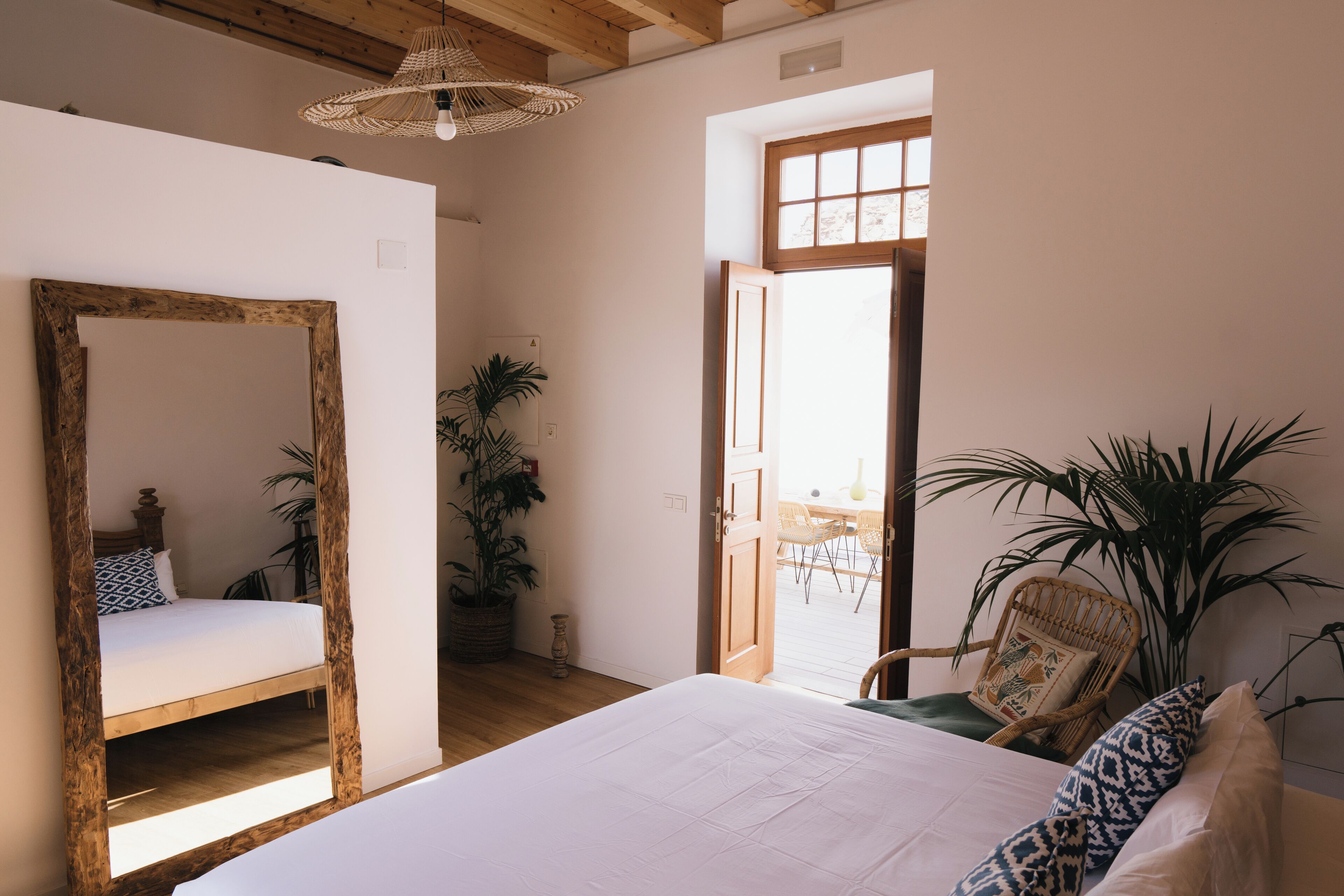 A bedroom with a large bed, a wooden-framed mirror, a potted plant, and a wicker chair.