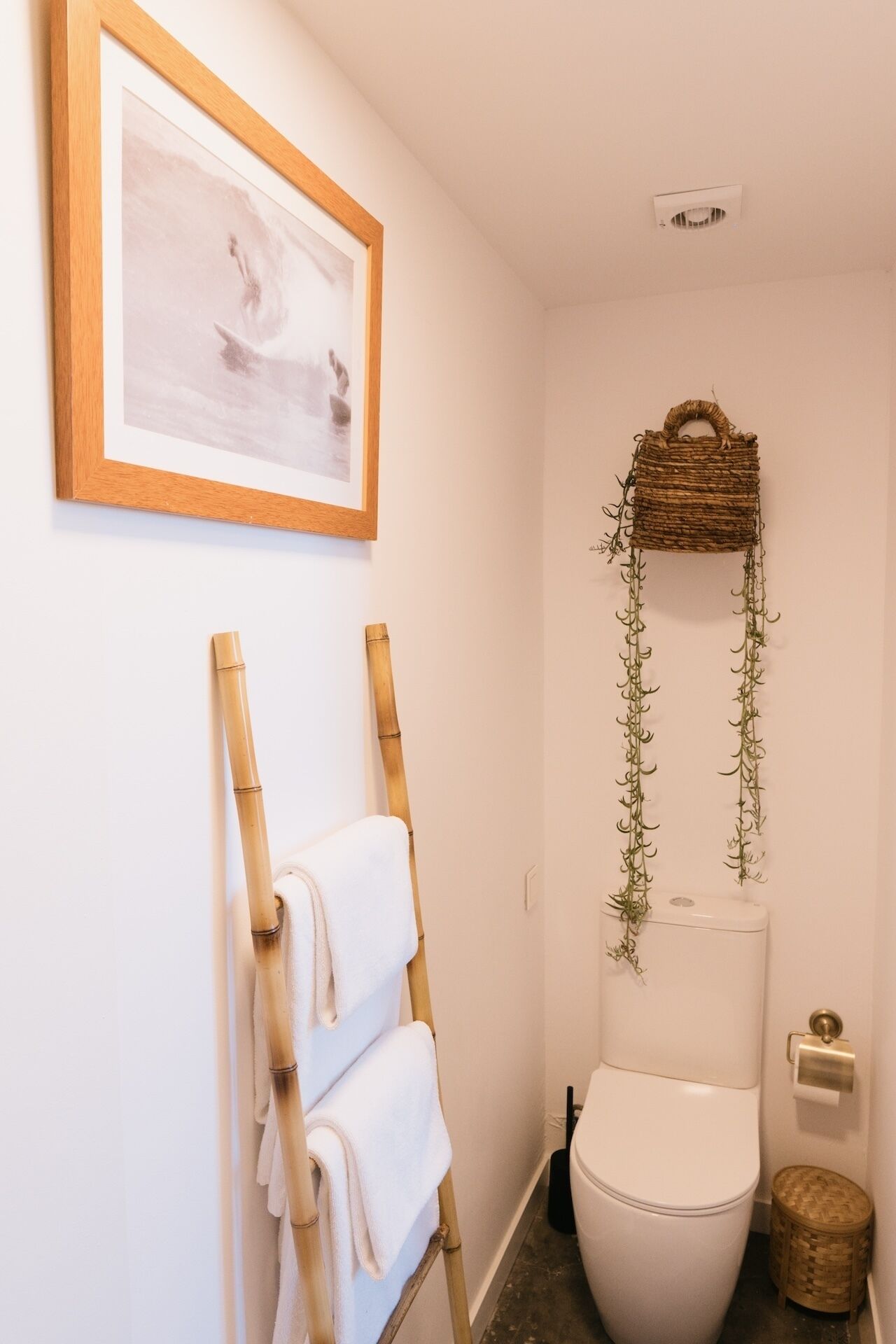 A bathroom with a toilet, a framed picture on the wall, a towel rack with towels, and a decorative hanging with greenery.