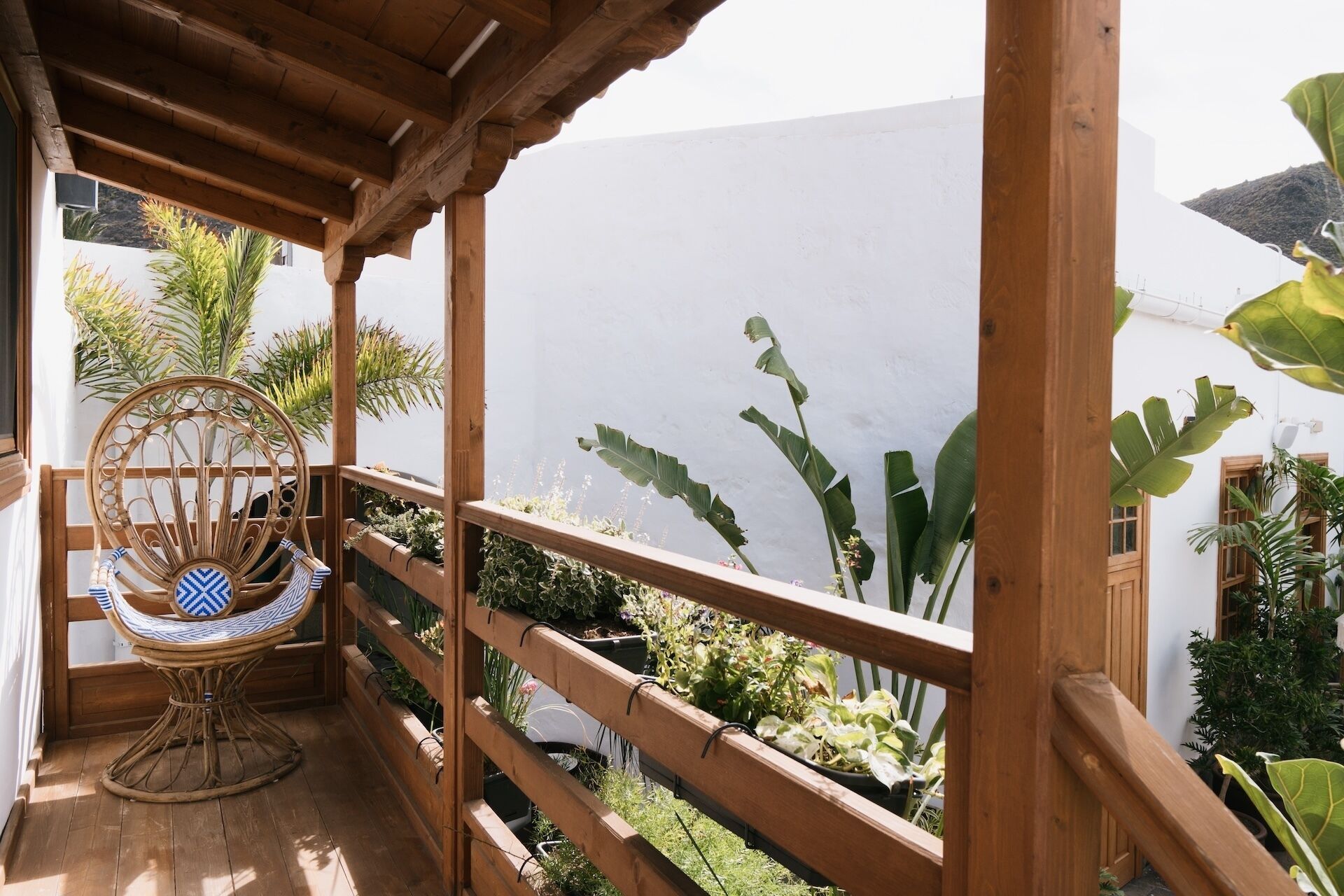Un balcón de madera con una silla de mimbre, plantas en macetas y vistas a un edificio blanco y montañas.