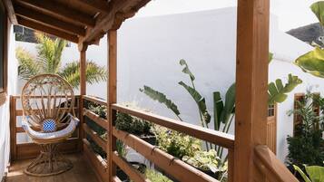 A wooden balcony with a wicker chair, potted plants, and a view of a white building and mountains.