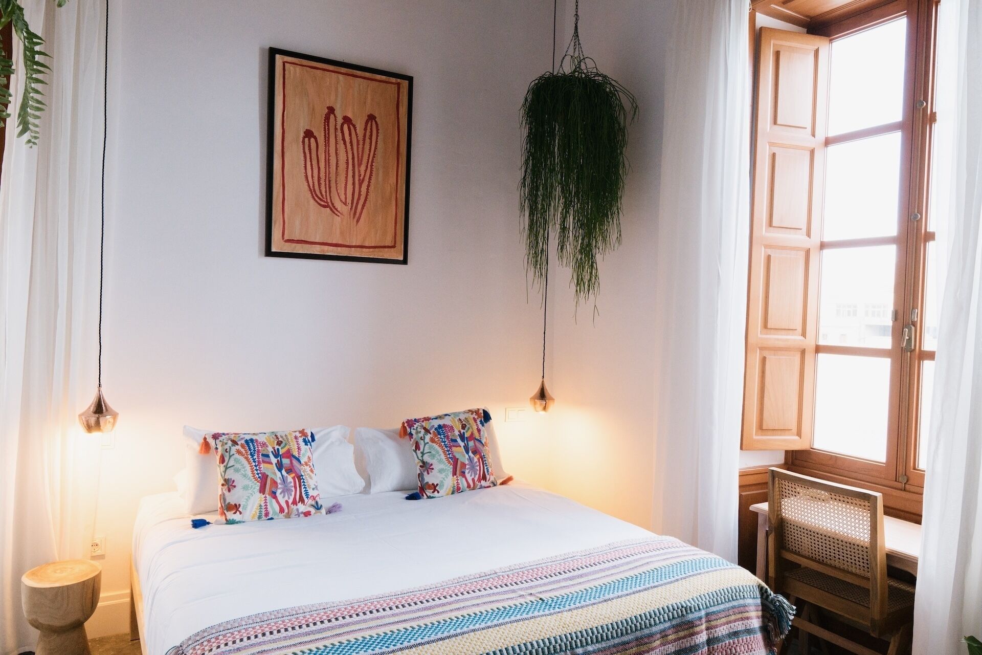 A bedroom with a bed, a hanging plant, a framed artwork, and a window with curtains.