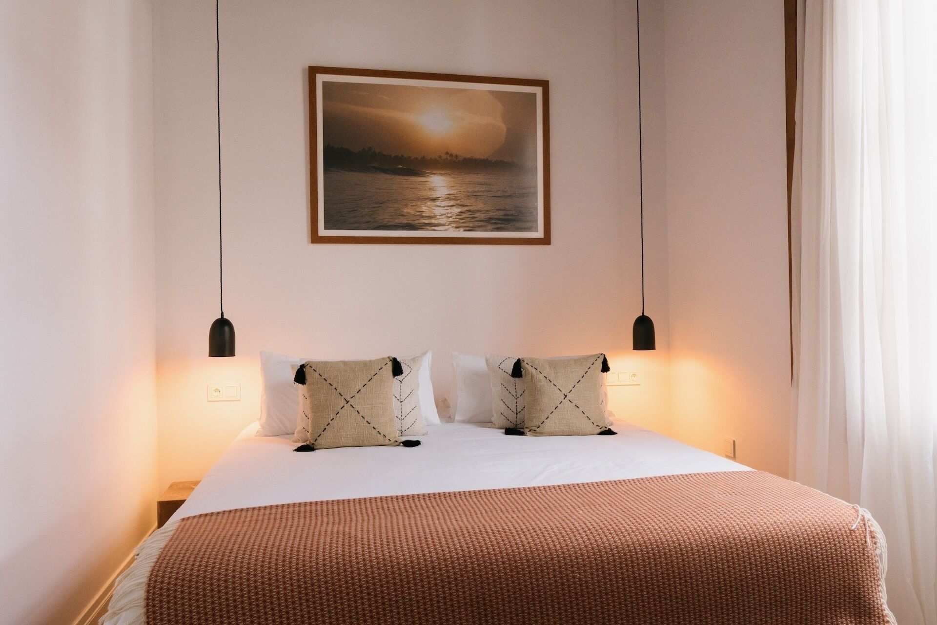 Una cama bien hecha con una manta marrón, dos almohadas blancas con borlas negras y un cuadro colgado en la pared que muestra un atardecer sobre el agua.