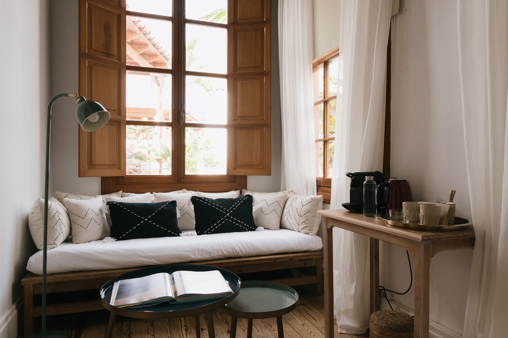 Ein gemütliches Zimmer mit einer hölzernen Bank, einem kleinen Tisch mit einem Buch, einem Fenster mit Vorhängen und einer Kaffeemaschine auf einem Beistelltisch.