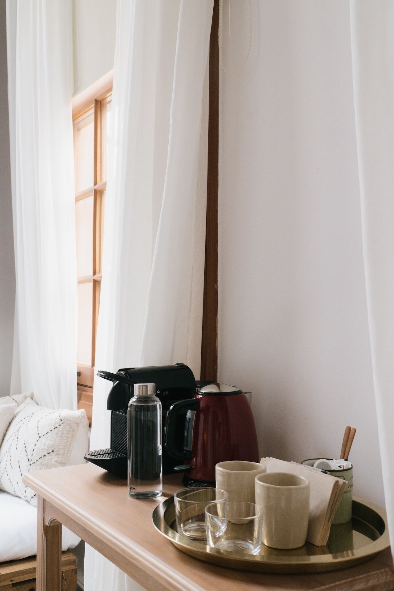 A breakfast setup with a coffee maker, a kettle, and a tray with cups and glasses on a wooden table near a window with sheer curtains.