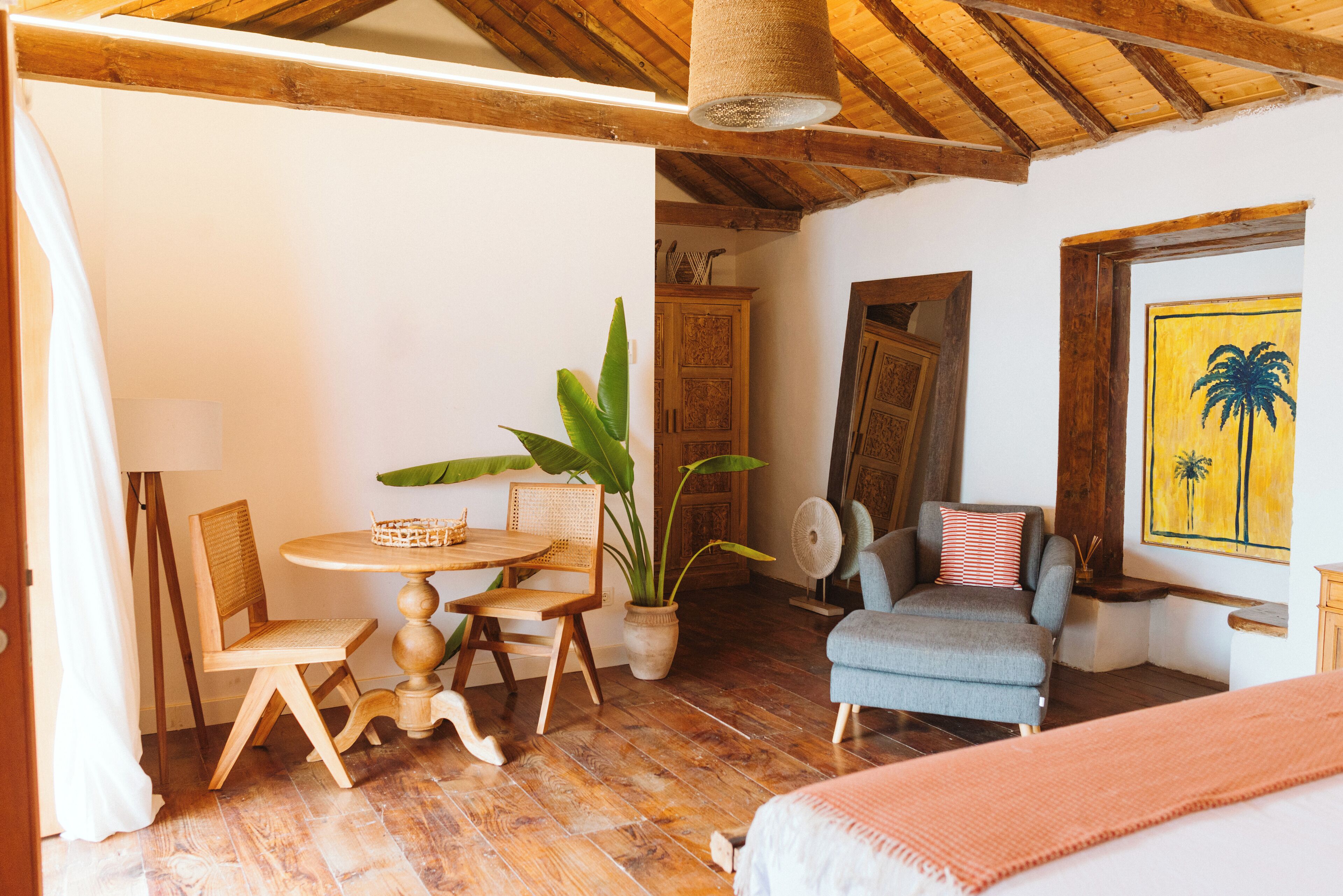 A cozy room with a wooden ceiling, a round wooden table with chairs, a blue armchair, a bed, and a large potted plant.