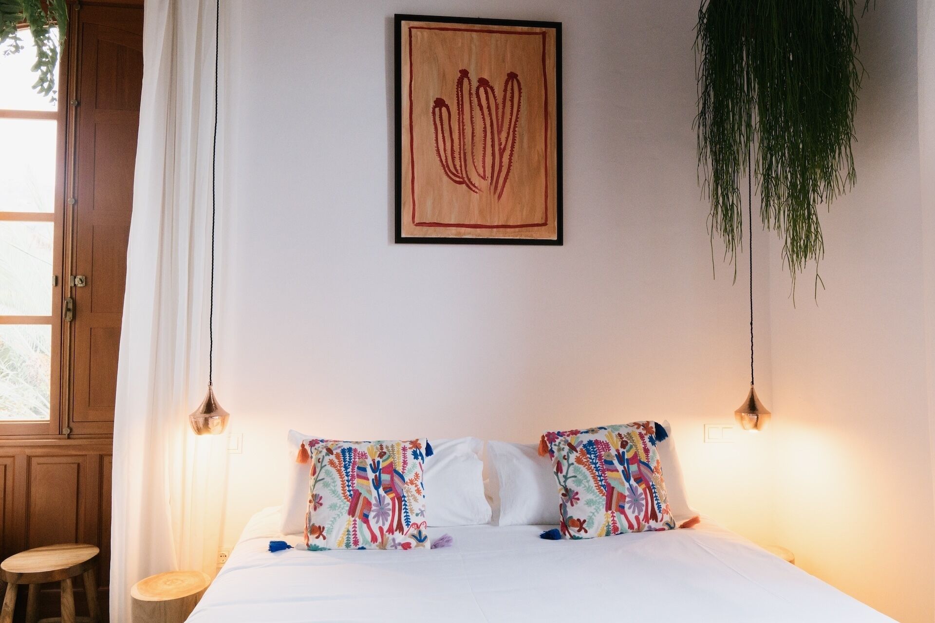 Un dormitorio con una cama, dos almohadas, una planta colgante, una obra de arte enmarcada y una ventana con cortinas.