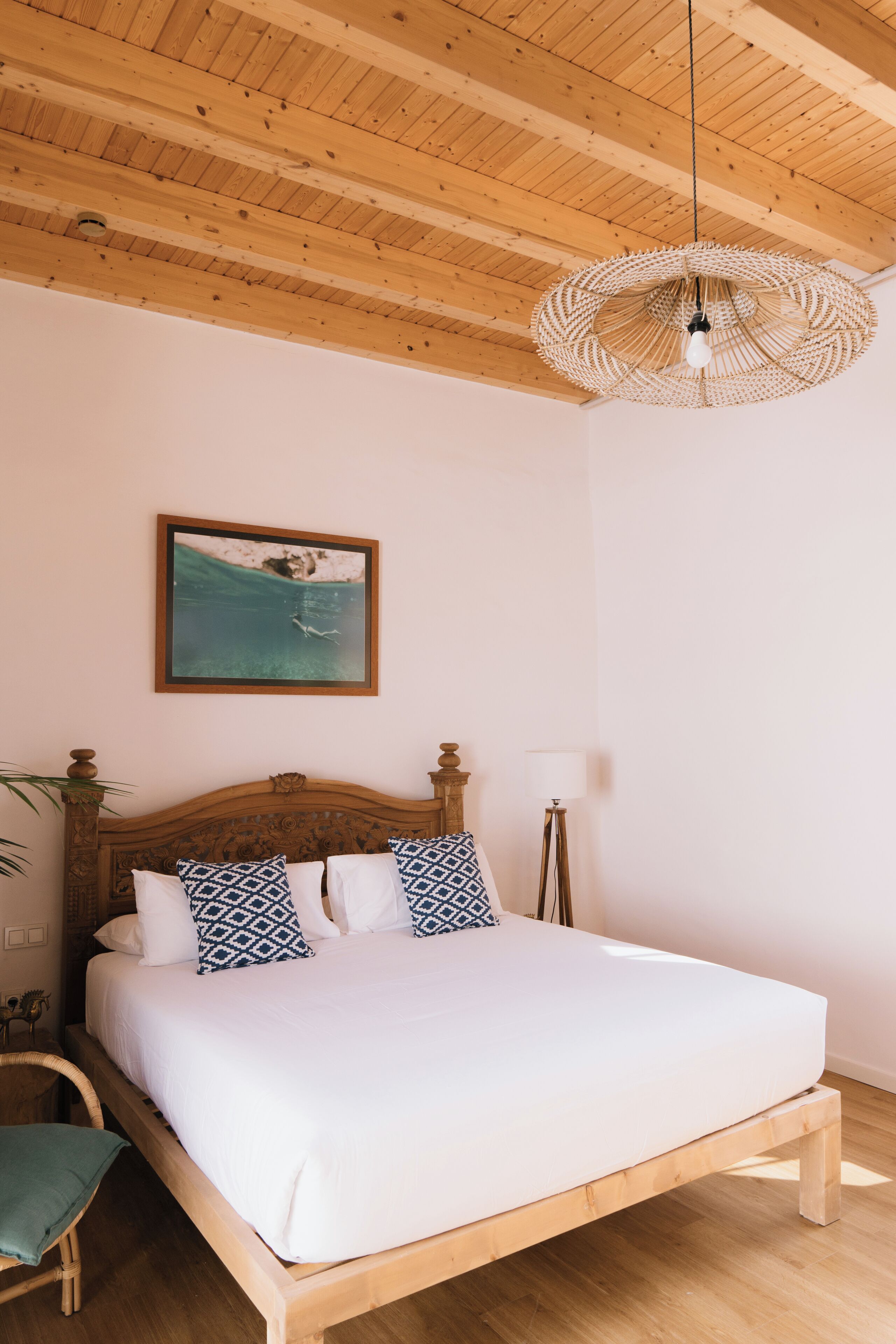 A bedroom with a wooden bed, a hanging light fixture, a framed picture on the wall, and a bedside table with a lamp.