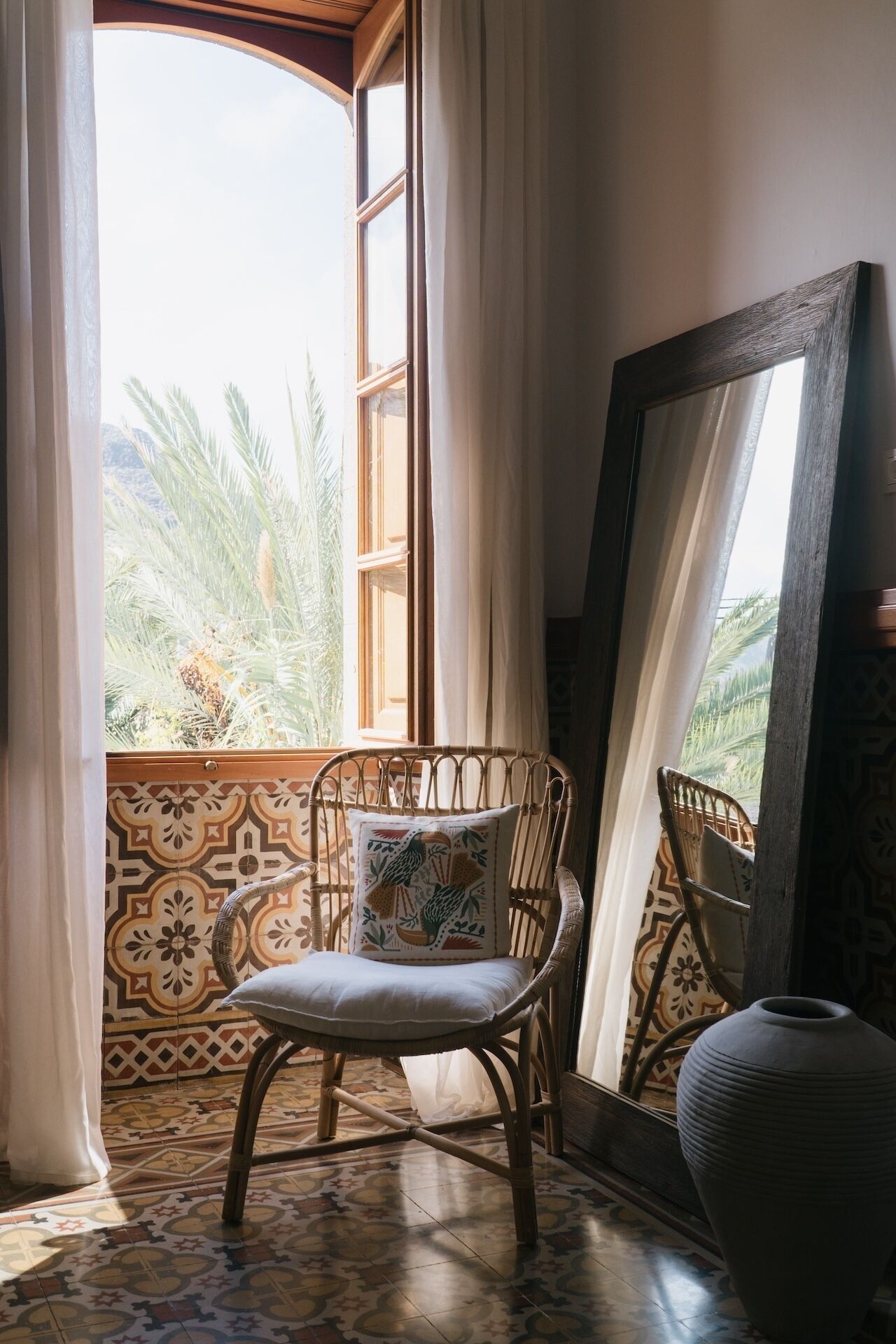 A wicker chair with a cushion and a decorative pillow, positioned near a window with curtains, overlooking a patterned floor and a view of palm trees.