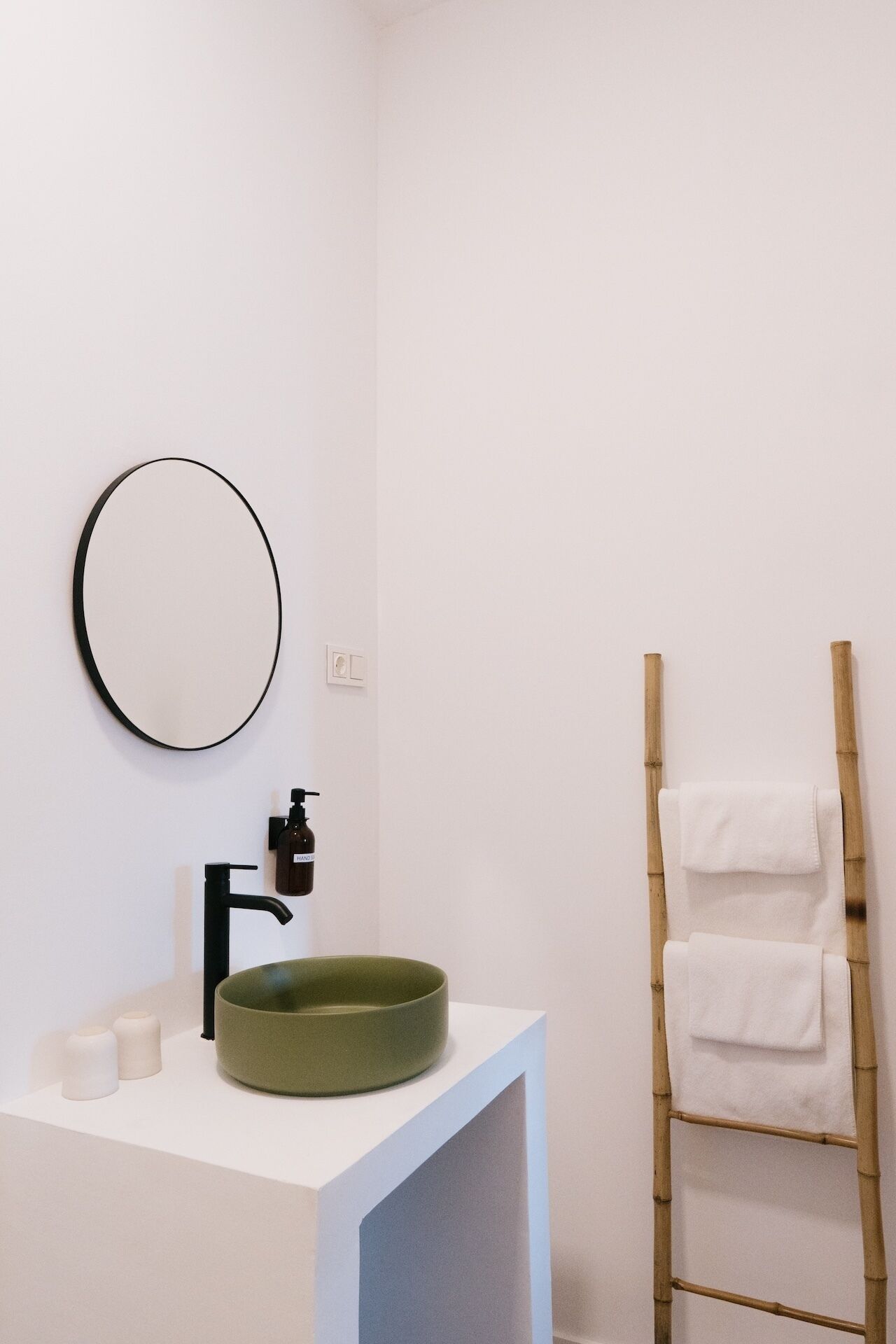 A bathroom with a white sink, a round mirror, a black faucet, and a bamboo ladder with towels.