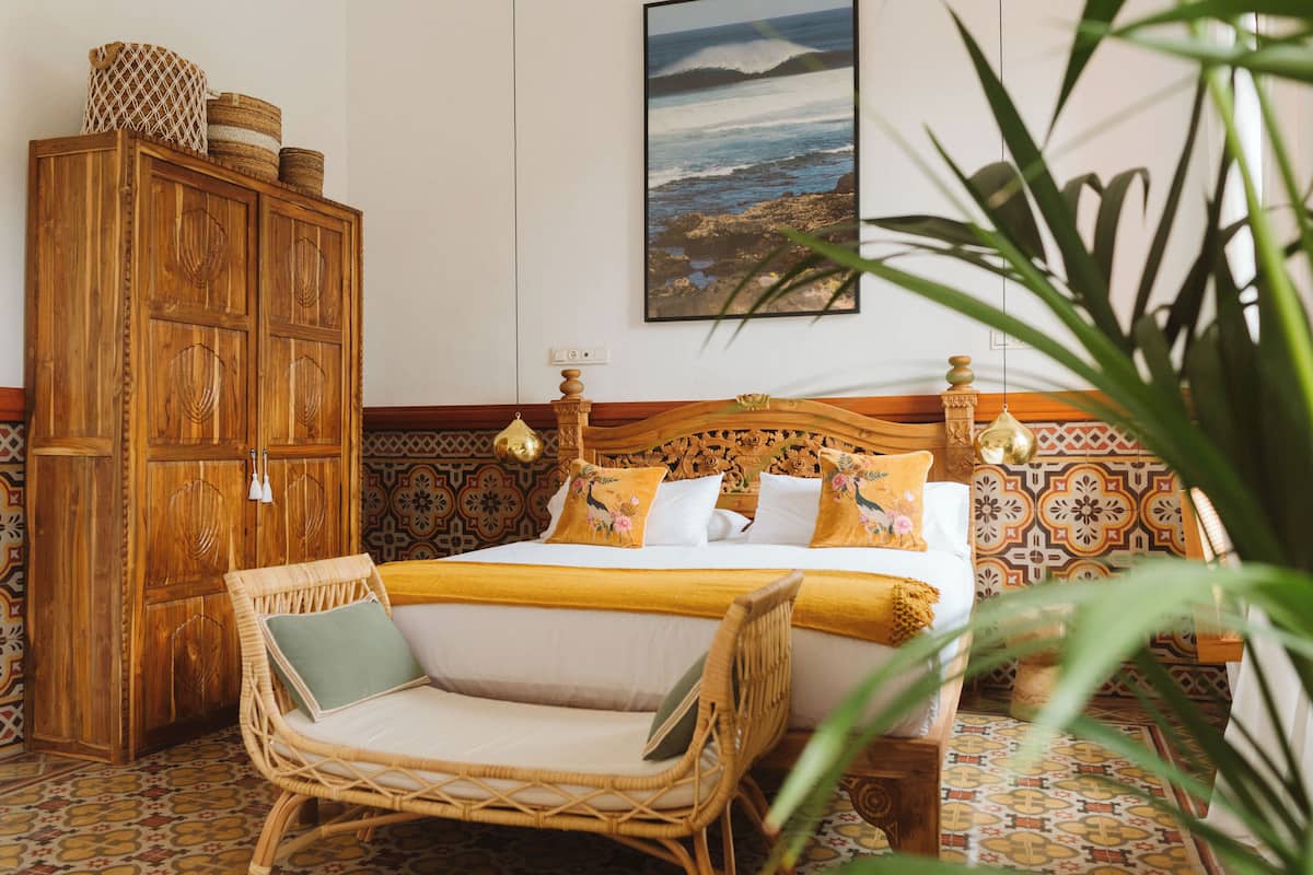 A bedroom with a wooden wardrobe, a bed with a patterned headboard, and a wicker chair.