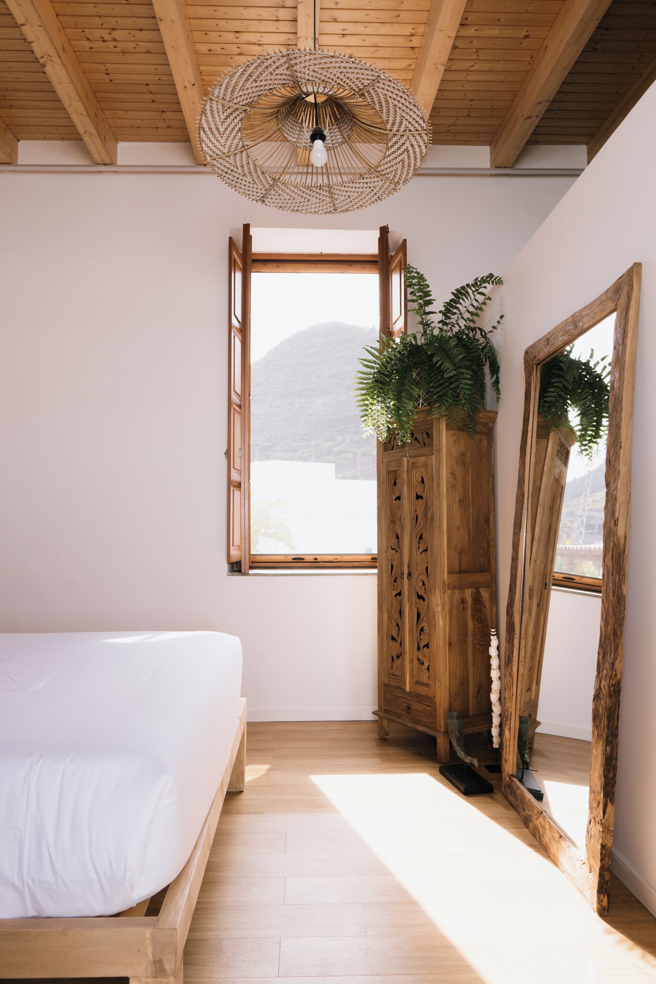 A bedroom with a wooden bed, a large window, a wooden wardrobe, and a mirror.
